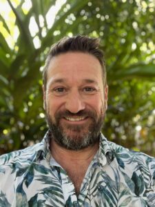 Photo of Byron Miller wearing a white and green shirt taken in 2023 at his home in Palmwoods on the Sunshine Coast Qld with some green plants in the background.
